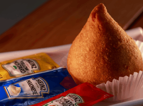 Coxinha dourada colocada em forminha de papel branco sobre bandeja, acompanhada de sachês de maionese, mostarda e ketchup da marca Heinz.