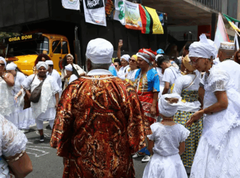 Grupo de pessoas vestidas com roupas tradicionais de matriz africana participa de celebração ao ar livre; muitas usam roupas brancas, turbantes e colares, enquanto outras vestem trajes coloridos. Banners e um caminhão de som aparecem ao fundo.