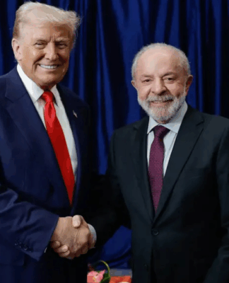 Dois homens de terno posam sorrindo enquanto se cumprimentam com um aperto de mão. Atrás deles aparecem as bandeiras dos Estados Unidos e do Brasil, com cortinas azuis ao fundo. À esquerda está o presidente americano, Donald Trump, e, à direita, o presidente brasileiro, Lula da Silva.