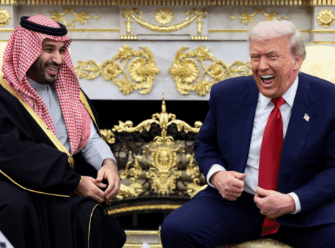 Dois homens sentados em poltronas claras conversam e riem em uma sala ornamentada com detalhes dourados; um deles veste terno azul com gravata vermelha, e o outro usa túnica tradicional preta com detalhes dourados e um lenço estampado vermelho e branco na cabeça.