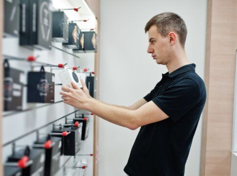 Homem branco e loiro de camiseta preta analisando produtos a venda em uma loja de eletrônicos