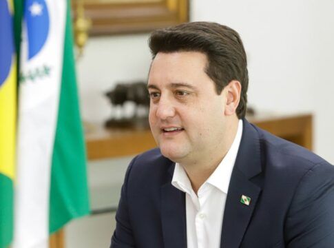 Ratinho Junior sentado em seu gabinete, com bandeira do Paraná ao fundo