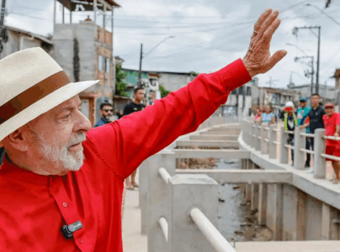 Homem de camisa vermelha e chapéu de palha acena sorridente em uma rua com obras e casas simples ao fundo, enquanto várias pessoas observam e registram o momento.