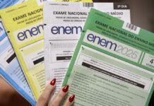 Mão segurando ps quatro cadernos de perguntas do ENEM. Um azul, um amarelo, um branco e um verde.