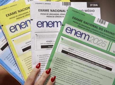 Mão segurando ps quatro cadernos de perguntas do ENEM. Um azul, um amarelo, um branco e um verde.
