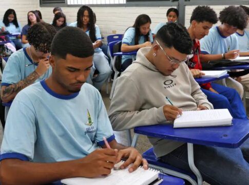 Foto de uma sala de aula. No plano frontal, um aluno negro e um aluno pardo fazendo anotações em seus cadernos