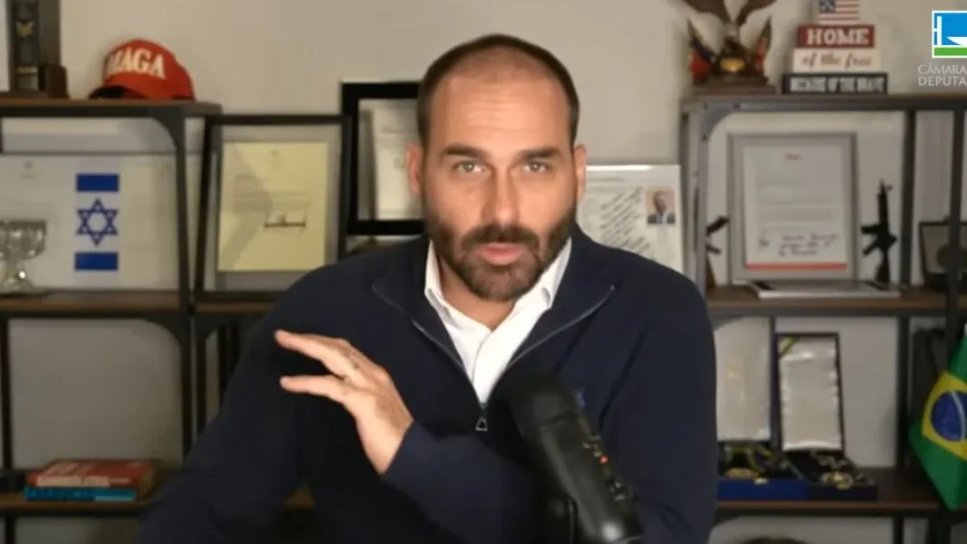 Eduardo Bolsonaro participando remotamente de uma sessão plenária. Ele está sentado à mesa com um microfone cardioide ao seu lado