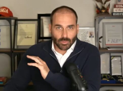 Eduardo Bolsonaro participando remotamente de uma sessão plenária. Ele está sentado à mesa com um microfone cardioide ao seu lado