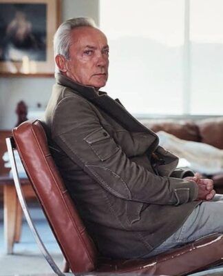 Ator Udo Kier veste um casaco e está sentado numa poltrona olhando de lado