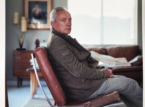 Ator Udo Kier veste um casaco e está sentado numa poltrona olhando de lado