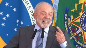 Presidente Lula, durante seu discurso, sorrindo e apontando com o dedo indicador para si mesmo. Ao fundo estão a bandeira do Brasil e cortinas azuis