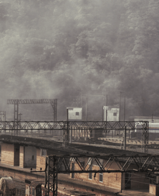 Paisagem da estação ferroviária de Paranapiacaba, com torre do relógio em destaque e trilhos ao redor. A cena está envolta por névoa densa, com vagões e trens visíveis diante de uma encosta coberta por mata.