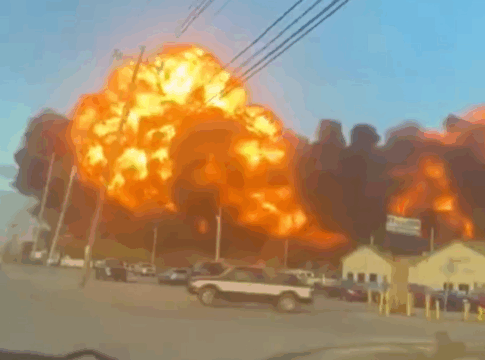Grande explosão em área industrial, com uma bola de fogo laranja e densa fumaça preta se espalhando pelo céu, enquanto carros estão estacionados e algumas construções aparecem em primeiro plano.