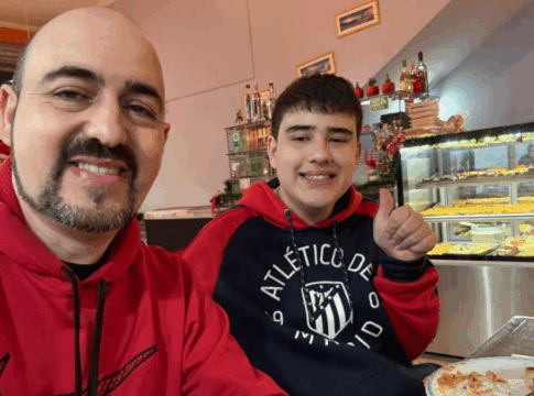Dois homens sorridentes sentados em uma mesa de restaurante posam para a foto; ambos usam moletom vermelho, um deles com o logotipo da Nike e o outro do Atlético de Madrid, enquanto fazem sinal de positivo com o polegar. Um prato com restos de pizza está à frente, e ao fundo há um balcão com vitrine de salgados e doces.