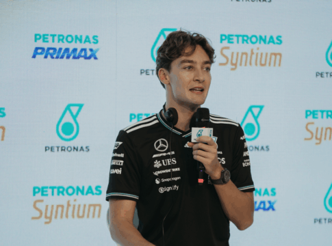 Homem veste camiseta preta da equipe Mercedes-AMG Petronas e segura um microfone durante entrevista ou coletiva de imprensa, com logotipos da Petronas e Syntium exibidos em um painel de fundo.
