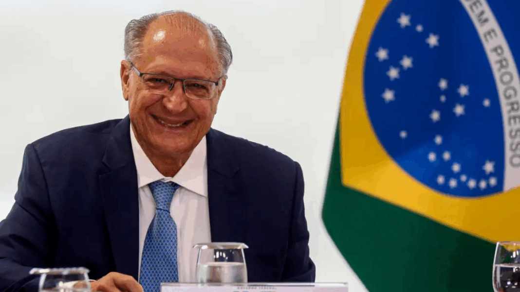 Homem de terno azul e gravata clara sorri sentado à mesa durante uma reunião oficial; à direita aparece parcialmente a bandeira do Brasil ao fundo, junto a copos de água e documentos sobre a mesa.