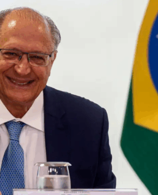 Homem de terno azul e gravata clara sorri sentado à mesa durante uma reunião oficial; à direita aparece parcialmente a bandeira do Brasil ao fundo, junto a copos de água e documentos sobre a mesa.