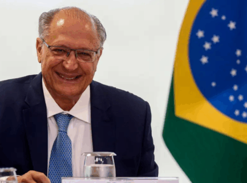 Homem de terno azul e gravata clara sorri sentado à mesa durante uma reunião oficial; à direita aparece parcialmente a bandeira do Brasil ao fundo, junto a copos de água e documentos sobre a mesa.