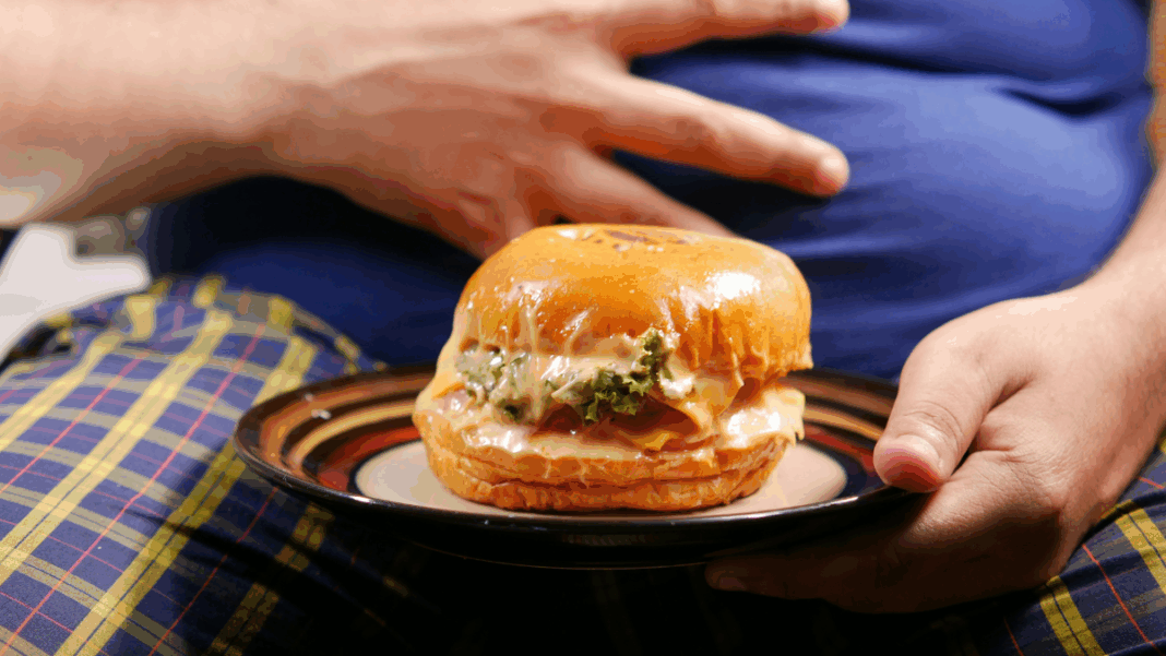 Pessoa sentada segura um prato com um hambúrguer grande e suculento; a mão repousa sobre a barriga coberta por camiseta azul, enquanto o sanduíche aparece em destaque à frente.