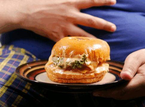 Pessoa sentada segura um prato com um hambúrguer grande e suculento; a mão repousa sobre a barriga coberta por camiseta azul, enquanto o sanduíche aparece em destaque à frente.