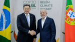 Dois homens de terno sorriem e se cumprimentam com um aperto de mão diante de bandeiras do Brasil e de Portugal, com o painel ao fundo exibindo o texto “COP30 Brasil Amazônia 2025 – Belém Climate Summit”.