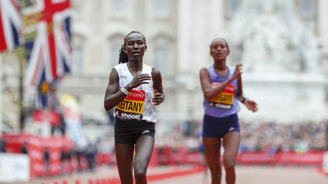 Duas atletas correm próximas à linha de chegada em uma maratona de rua. A corredora à frente, usando camiseta branca e tênis rosa, exibe expressão de esforço. Bandeiras do Reino Unido e o público aparecem ao fundo.