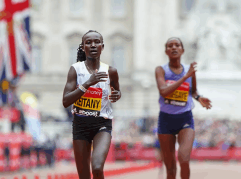 Duas atletas correm próximas à linha de chegada em uma maratona de rua. A corredora à frente, usando camiseta branca e tênis rosa, exibe expressão de esforço. Bandeiras do Reino Unido e o público aparecem ao fundo.