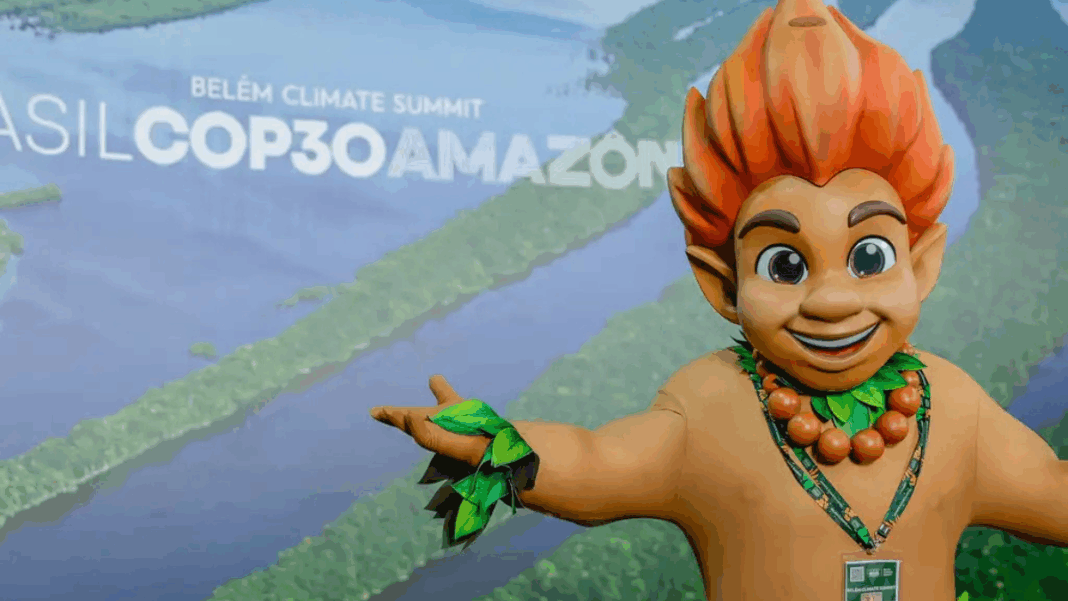 Mascote de aparência infantil, com cabelo laranja em forma de chamas e adereços de folhas verdes, posa sorridente com os braços abertos diante de painel com imagem aérea da Floresta Amazônica e o texto “Brasil COP30 Amazônia – Belém Climate Summit”.