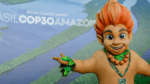 Mascote de aparência infantil, com cabelo laranja em forma de chamas e adereços de folhas verdes, posa sorridente com os braços abertos diante de painel com imagem aérea da Floresta Amazônica e o texto “Brasil COP30 Amazônia – Belém Climate Summit”.