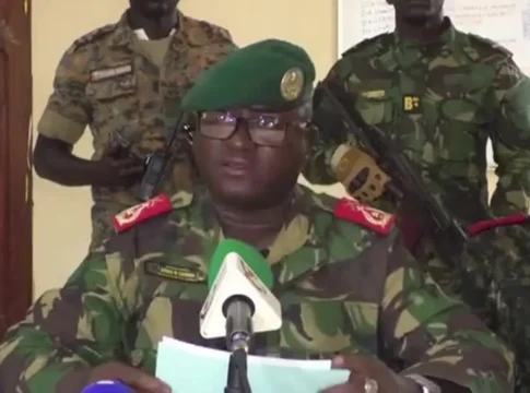 Oficial militar com boina verde e farda camuflada lê um comunicado diante de microfones, enquanto soldados armados permanecem em pé atrás dele dentro de uma sala administrativa.