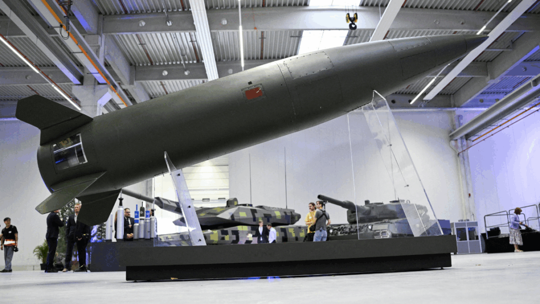 Míssil de grande porte exposto em um suporte transparente dentro de um pavilhão industrial, com tanques militares ao fundo e visitantes observando os equipamentos.