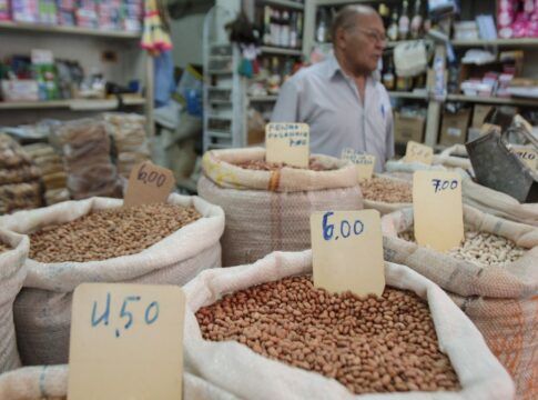 A imagem mostra uma banca de feira, com sacos de graõs e preços variados