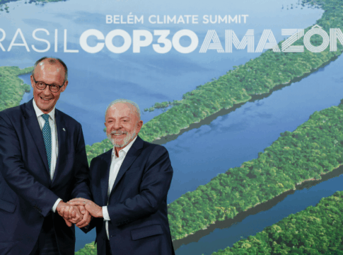 Dois homens de terno sorriem e apertam as mãos diante de um painel com imagem aérea da Floresta Amazônica e o texto “Brasil COP30 Amazônia – Belém Climate Summit”.