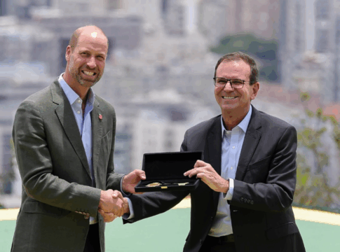 Dois homens de terno trocam um aperto de mão enquanto um deles segura uma caixa aberta com uma chave dourada em seu interior, com prédios desfocados ao fundo.