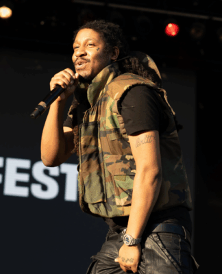 Homem segura um microfone enquanto canta em um palco durante um show ao ar livre. Ele veste colete camuflado e calça preta, com a palavra “SUMMERFEST” visível no fundo.