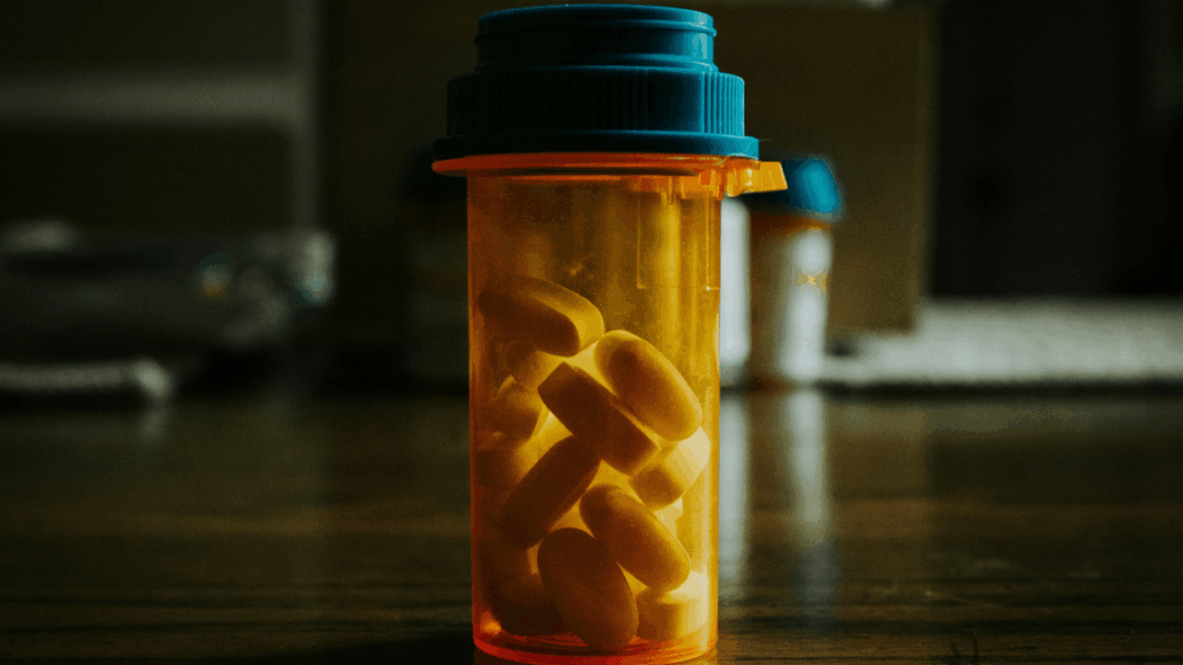 Frasco transparente com tampa azul contendo comprimidos amarelos sobre uma mesa de madeira, em ambiente interno com iluminação suave e fundo desfocado.