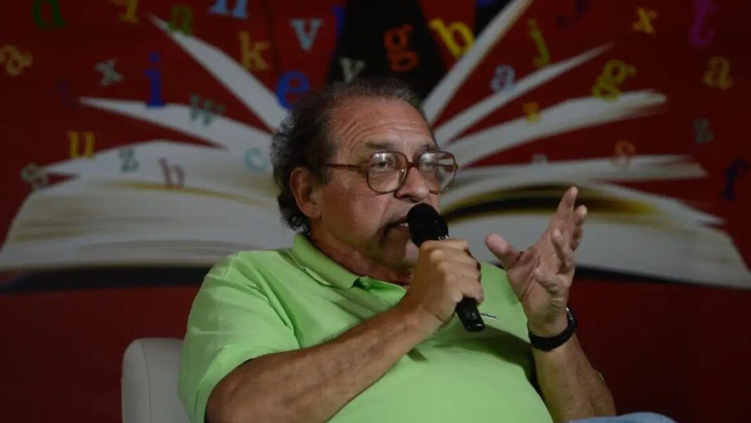 Rui Castro , vestindo camisa polo verde-clara, falando ao microfone durante um evento literário. Ao fundo, há uma imagem de um livro aberto com letras coloridas espalhadas sobre um painel vermelho