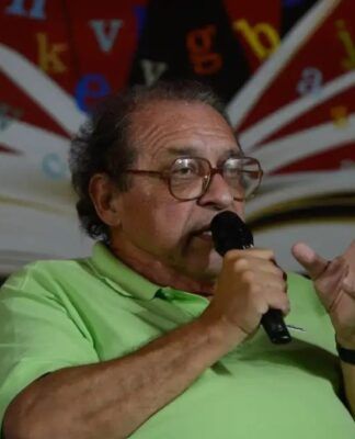 Rui Castro , vestindo camisa polo verde-clara, falando ao microfone durante um evento literário. Ao fundo, há uma imagem de um livro aberto com letras coloridas espalhadas sobre um painel vermelho