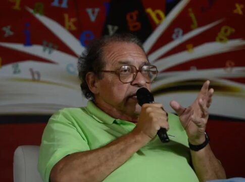 Rui Castro , vestindo camisa polo verde-clara, falando ao microfone durante um evento literário. Ao fundo, há uma imagem de um livro aberto com letras coloridas espalhadas sobre um painel vermelho