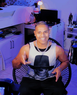 Homem sorri enquanto grava um vídeo em um quarto iluminado por luzes coloridas e equipamentos de gravação, sentado em frente a uma câmera e ring light, com prateleiras e objetos decorativos ao fundo.