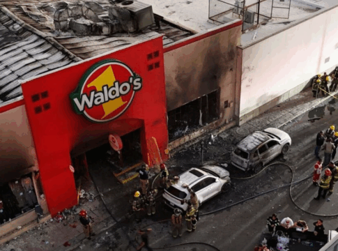 Imagem aérea mostra fachada queimada de uma loja Waldo’s, no México, após um incêndio. Bombeiros e equipes de resgate trabalham no local, ao lado de carros carbonizados e escombros espalhados pela rua.