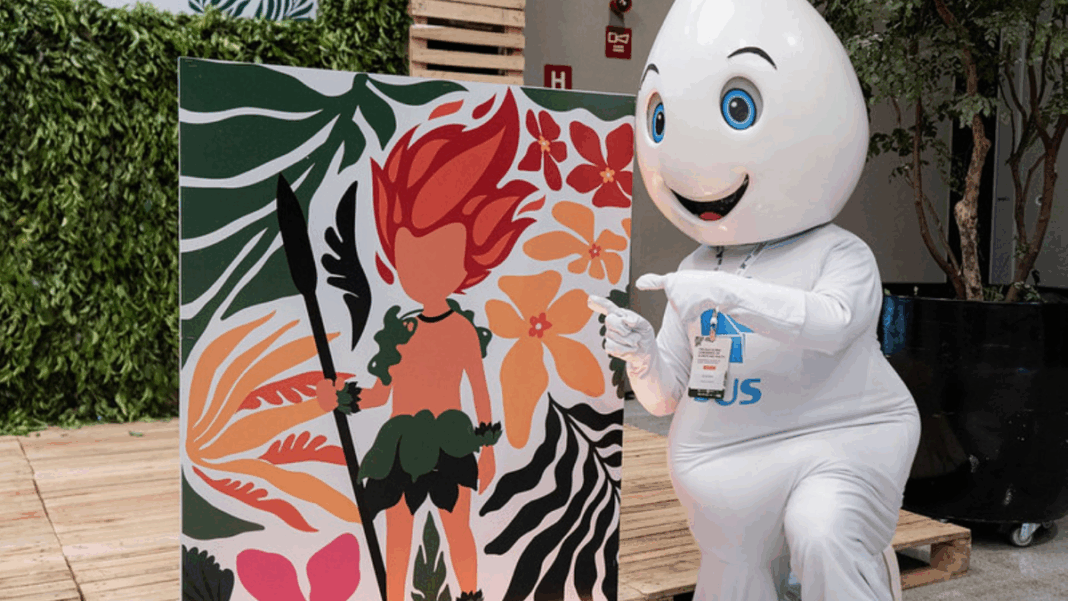 Mascote em formato de gota d’água, com olhos grandes e expressão sorridente, ajoelha-se ao lado de um painel colorido que mostra ilustração de personagem de cabelo vermelho em meio a folhas e flores tropicais, apontando para a arte.