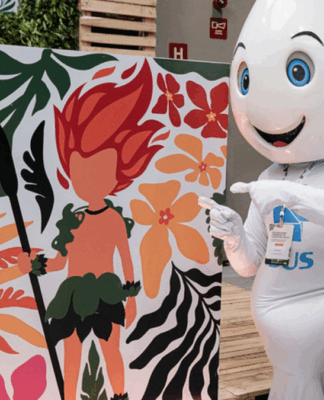 Mascote em formato de gota d’água, com olhos grandes e expressão sorridente, ajoelha-se ao lado de um painel colorido que mostra ilustração de personagem de cabelo vermelho em meio a folhas e flores tropicais, apontando para a arte.