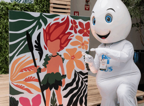 Mascote em formato de gota d’água, com olhos grandes e expressão sorridente, ajoelha-se ao lado de um painel colorido que mostra ilustração de personagem de cabelo vermelho em meio a folhas e flores tropicais, apontando para a arte.