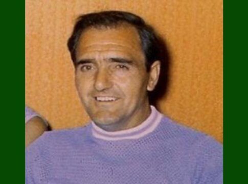 A foto é um retrato em close-up de um homem de meia-idade identificado como Alex Mitchell, da década de 1970. O homem tem cabelo escuro, curto e penteado de forma clássica. Ele parece ter uma tez clara e um sorriso aberto, que revela os dentes. As sobrancelhas são grossas e escuras.