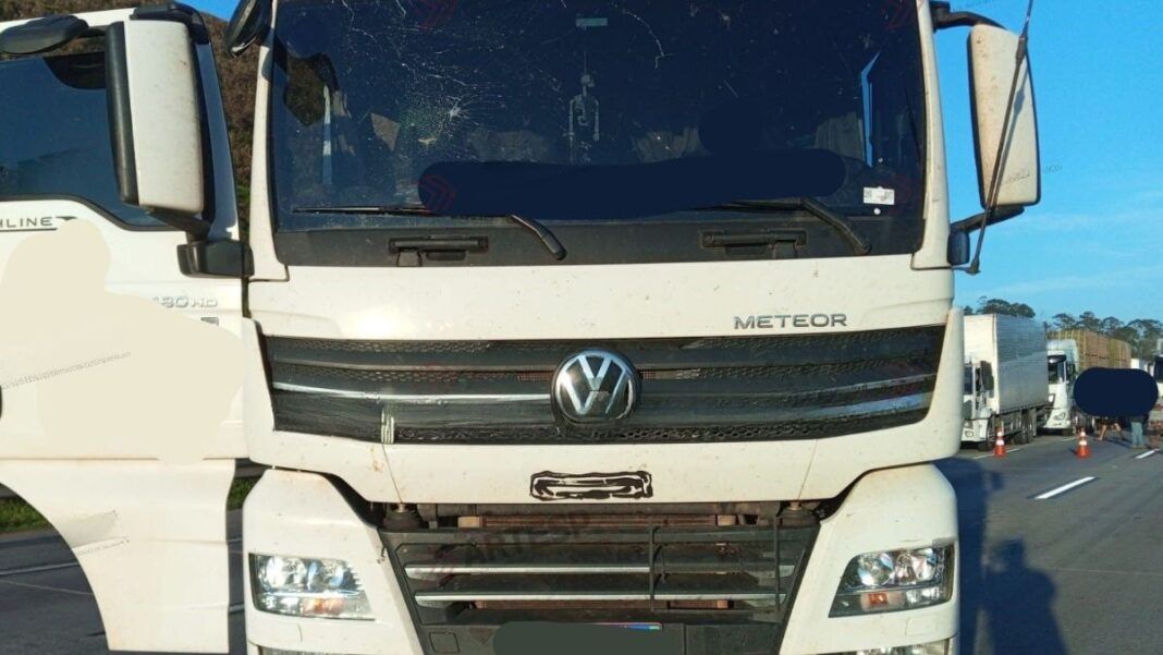 Imagem mostra caminhão branco parado no meio de uma rodovia. O para-brisa do veículo tem uma avaria, com um buraco no canto superior esquerdo