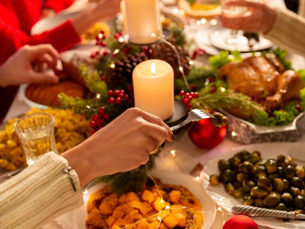 Na foto há uma mesa de jantar em que há muitos pratos, velas e pessoas comendo.