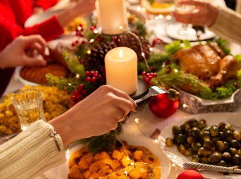 Na foto há uma mesa de jantar em que há muitos pratos, velas e pessoas comendo.