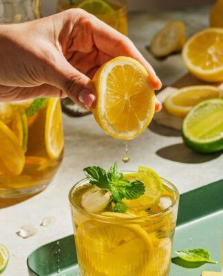 A imagem mostra uma pessoa espremendo uma laranja em um copo com chá que está em cima de uma bandeja verde