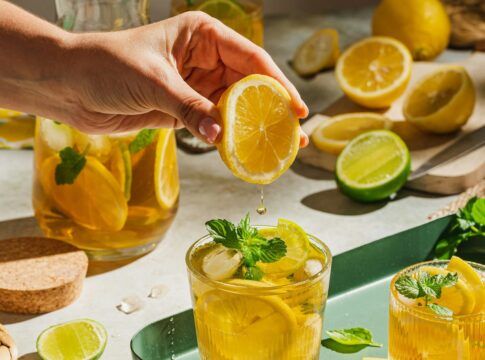 A imagem mostra uma pessoa espremendo uma laranja em um copo com chá que está em cima de uma bandeja verde